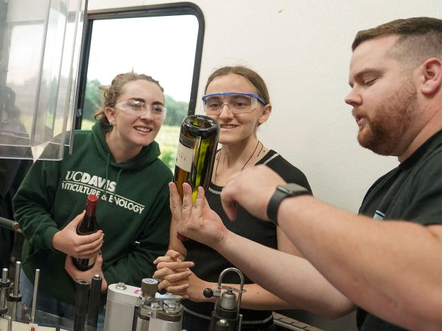 哈尔西装瓶厂的一名员工举起一个酒瓶并指着它，同时向两名戴着护目镜微笑的学生解释了一个要点。