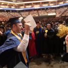 一名男性毕业生手举着手，大步走在两排教员之间，挥舞着彩球和拍子。 