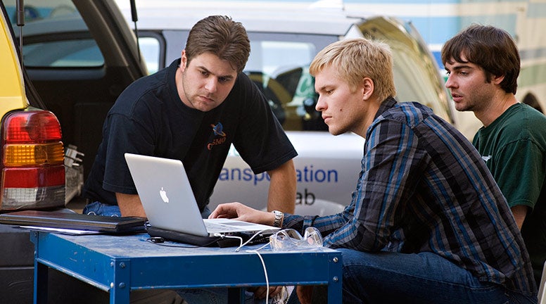  Three men hover over a computer with a car in the background