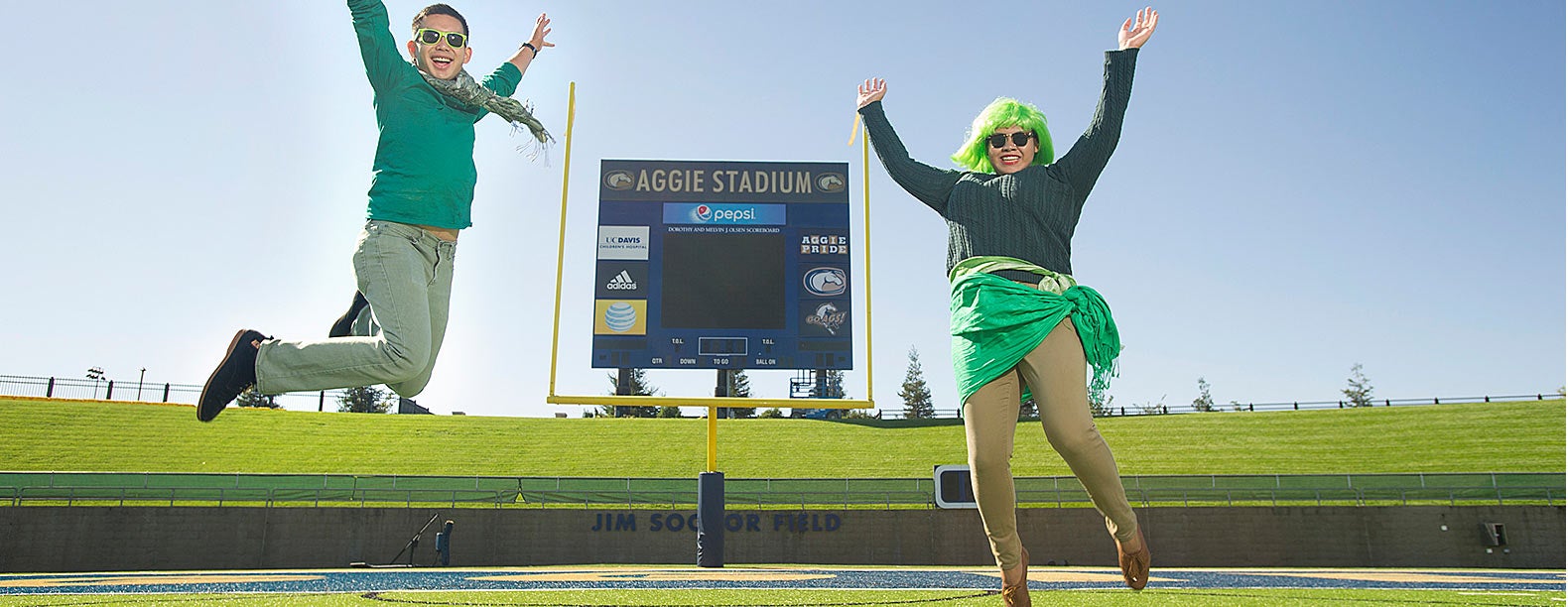  A woman and a man dressed in several pieces of green clothing pose in front of the Aggie Stadium scoreboard