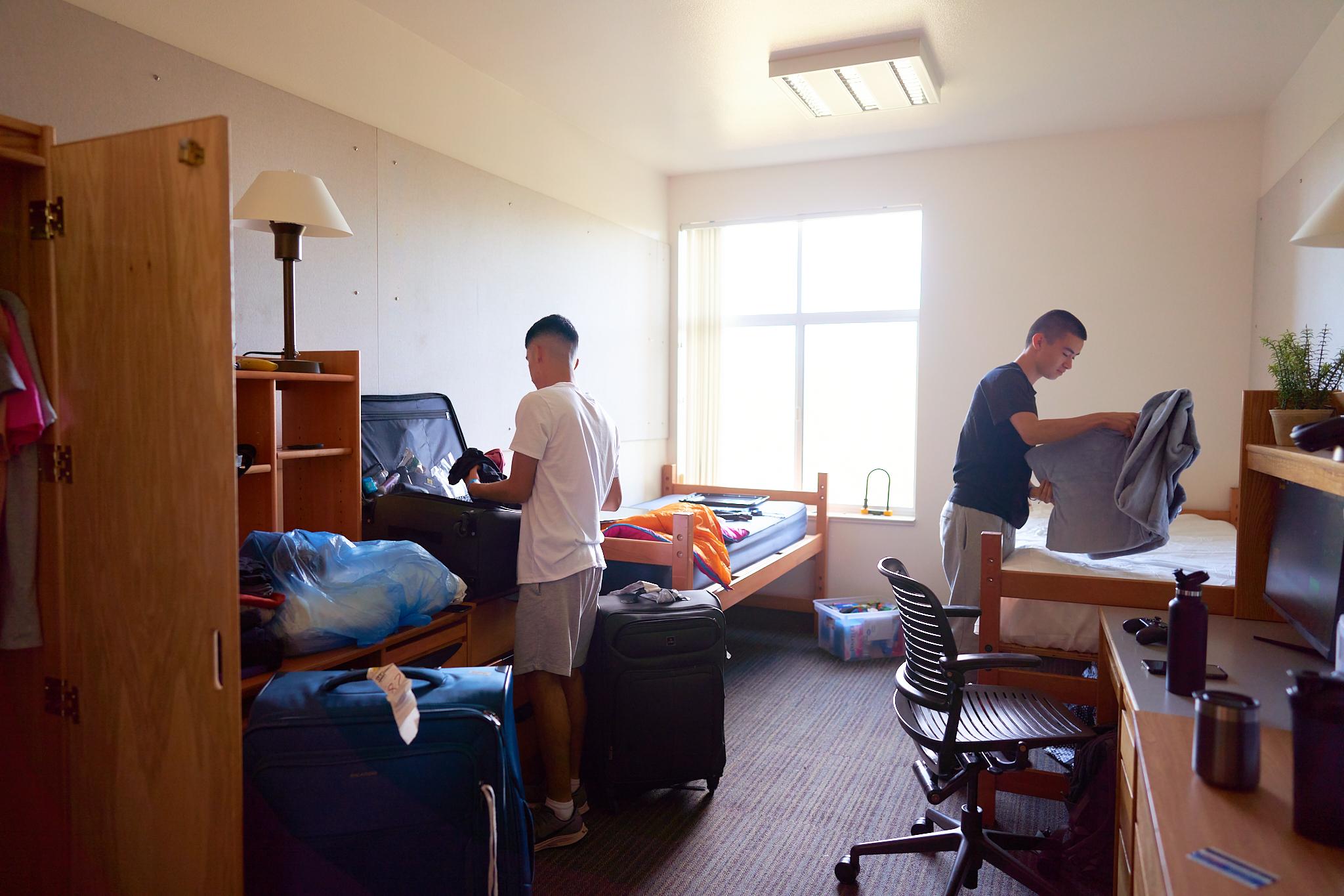 Two students in a UC Davis’ residential double dorm room unpack their belongings from suitcases and other bags. 