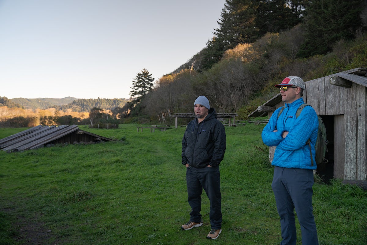 Barry McCovey in black coat and Rob Lusardi in Blue stand in grassy ceremonial area looking at Klamath River