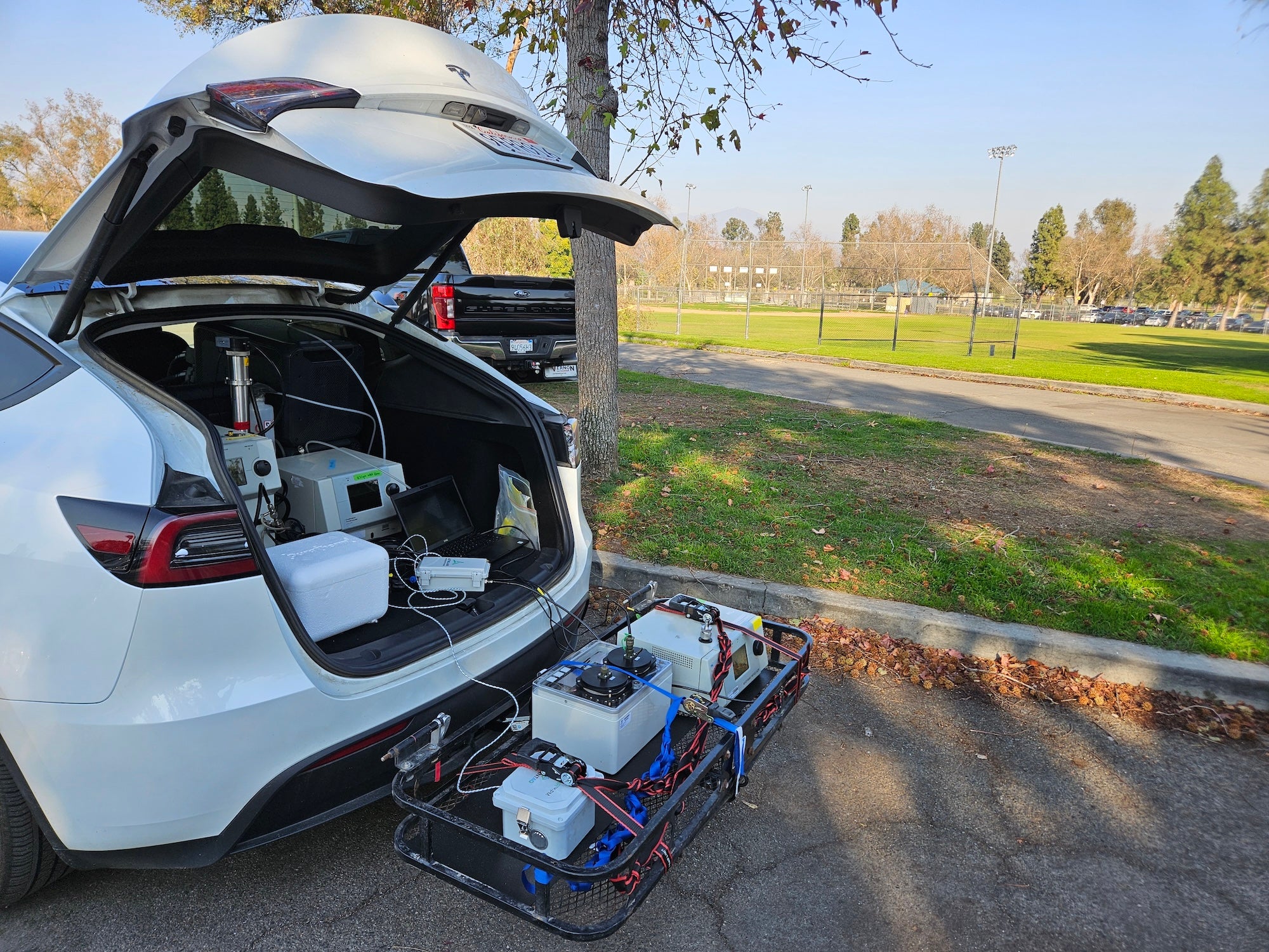 White car with open trunk holds air sampling and monitoring equipment. Background is green grass and tree. 