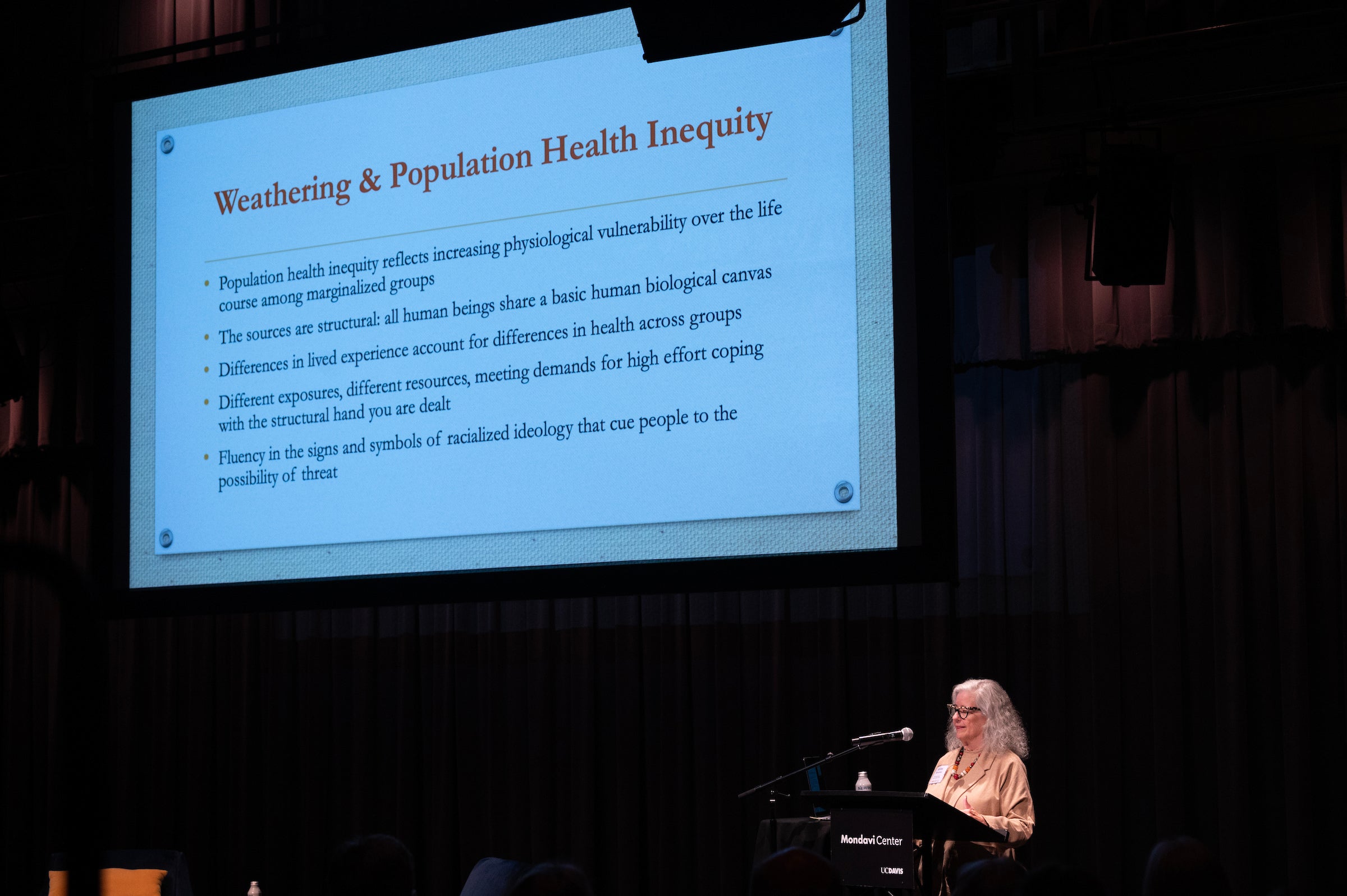 a large slide from a presentation is projected onto a theater's screen. In the bottom right corner stands Dr. Arline T. Geronimus in front of a lectern, speaking into a microphone. The presented screen defines the term "weathering" and its impact on population health inequity. 