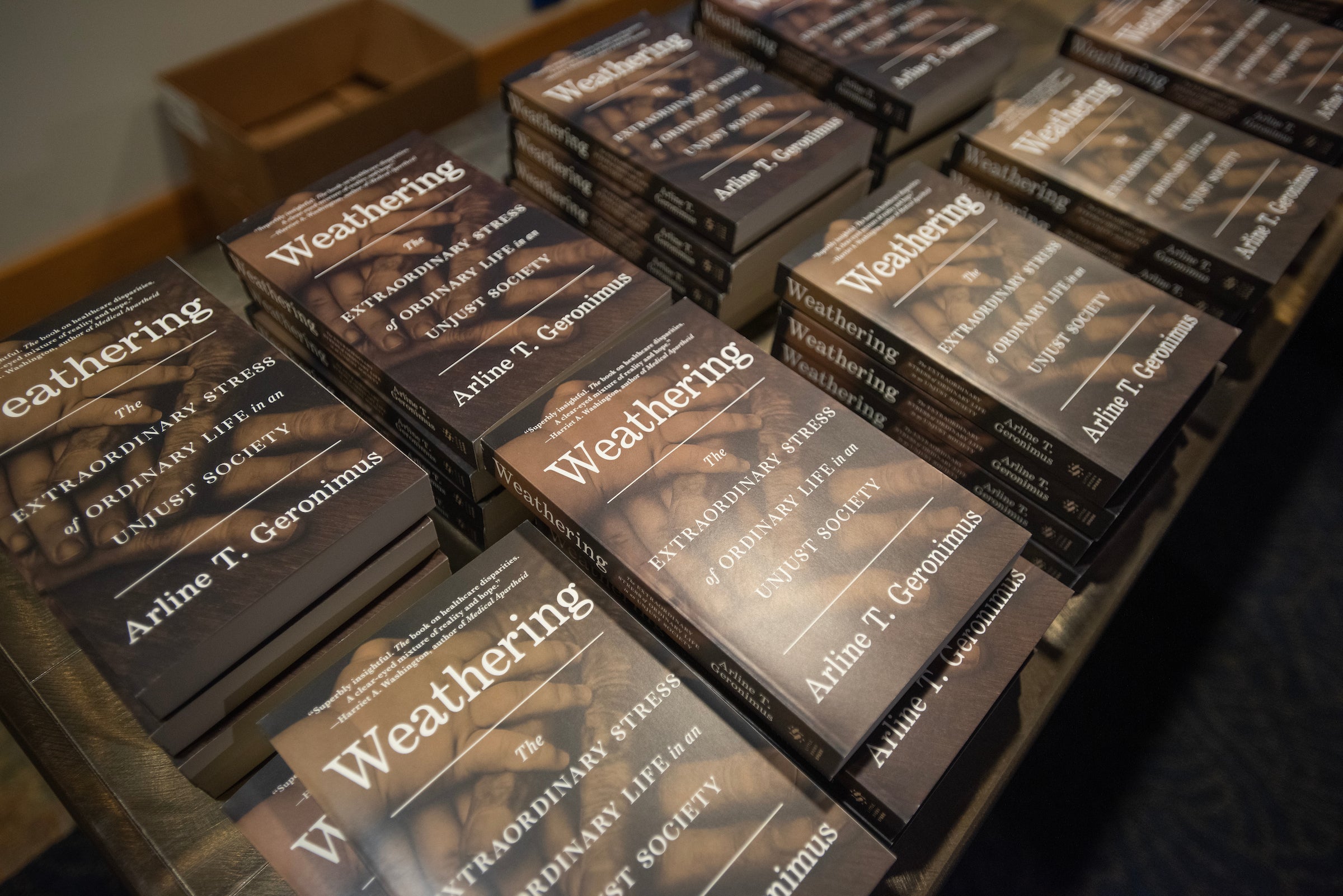 a Birds Eye view perspective of a table full of copies of the book "Weathering." The book cover is black with white text, and a silhouetted image of multiple hands of different skin tones, with their fingers extended, showcasing their skin.