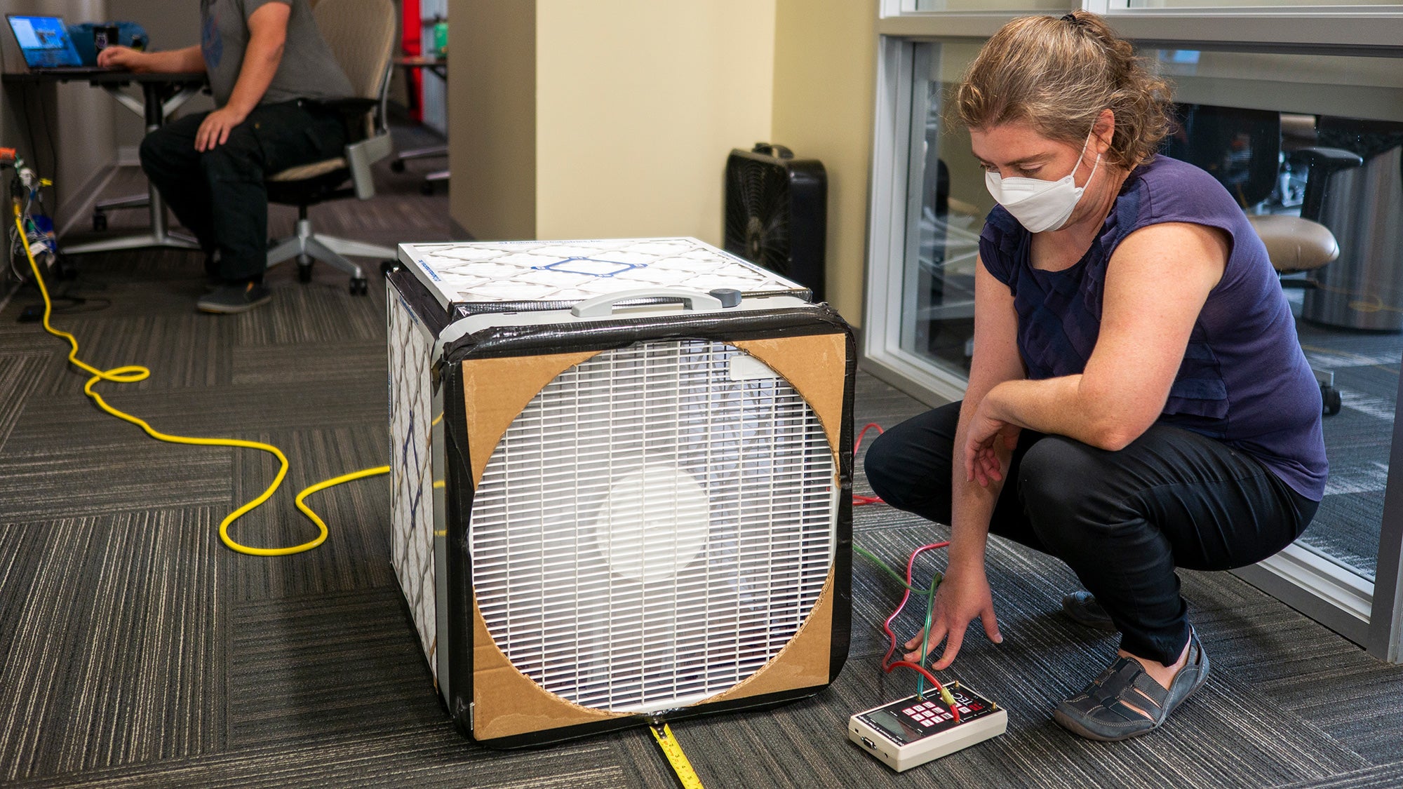 A woman kneels next to a box with air filters on it to monitor the air quality of a room