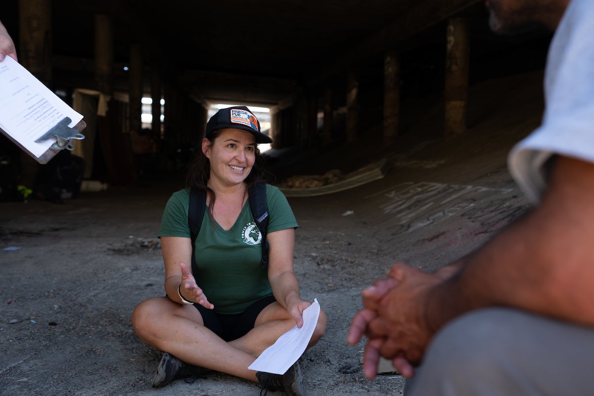 Costanza Rapini sits crosslegged on concrete floor talking with man whose arm and knee are visible