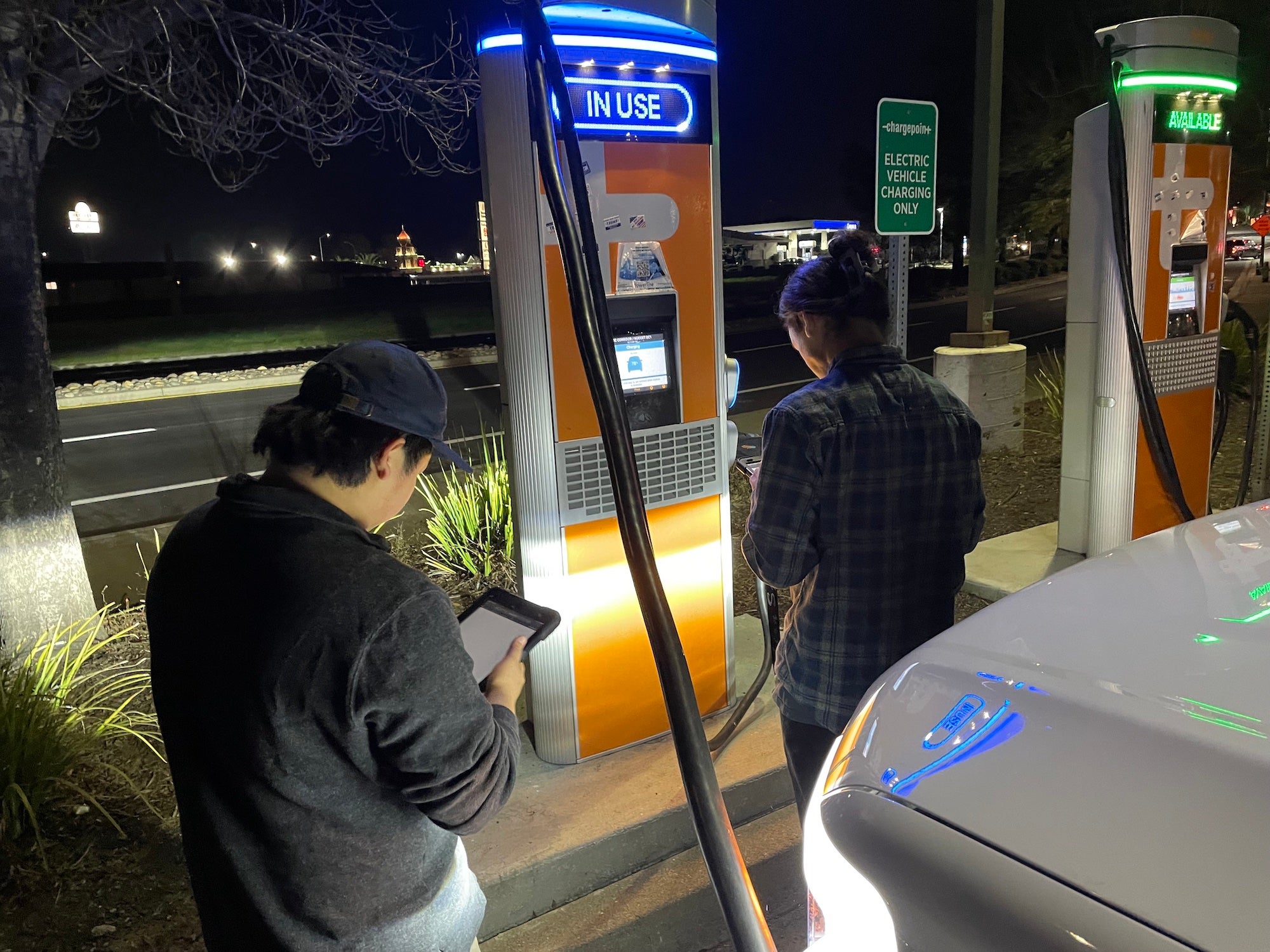 UC Davis students Joshua Bang, left, and Brian Olszewski, right, stand in front of a Charge Point electric vehicle charging station at night while charging a Ford F-150 Lightning. They are helping researchers test the reliability of thousands of electric vehicle charging stations across California. (Amy Quinton / UC Davis)