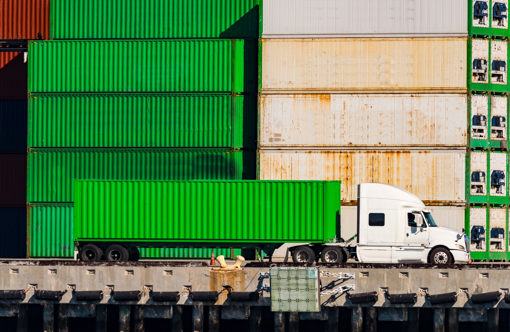 bright green heavy-duty truck sits parallel to stack of cargo containers at shipping port