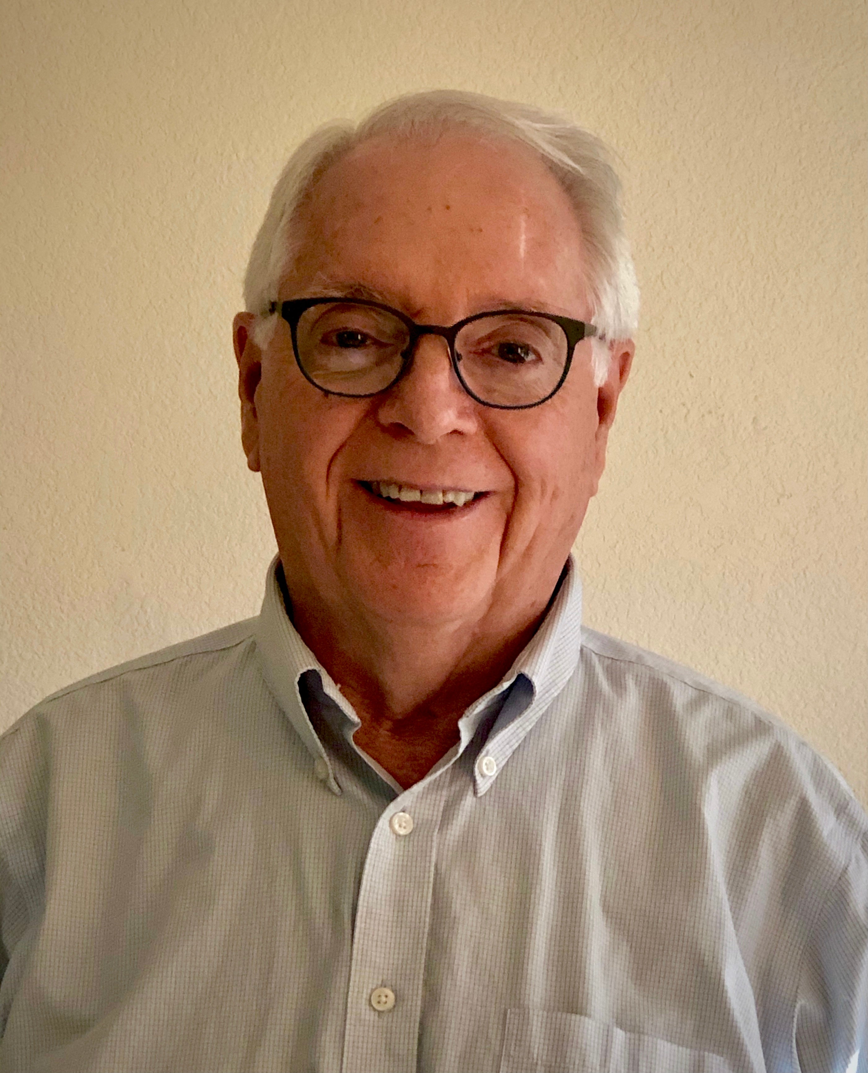 Distinguished Professor Michael Savageau is standing centered in front of the camera. He's smiling while wearing black rimmed glasses, a grey Oxford shirt and short white hair. 