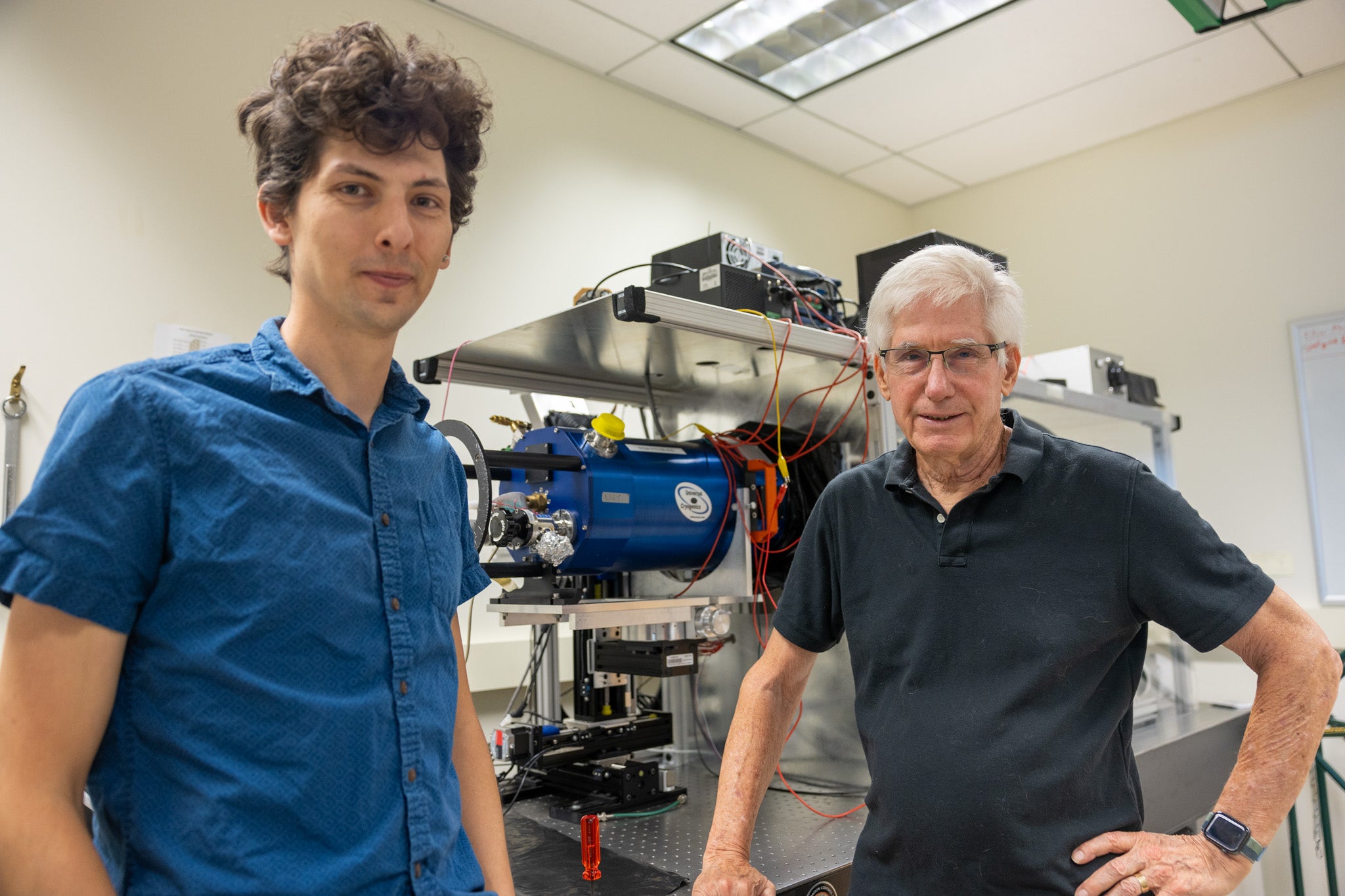 A young man with brown curly hair and a blue shirt and an older man with grey hair and a dark blue shirt look towards the camera in an indoor lab. Behind them is an apparatus with wires and cylinders. 