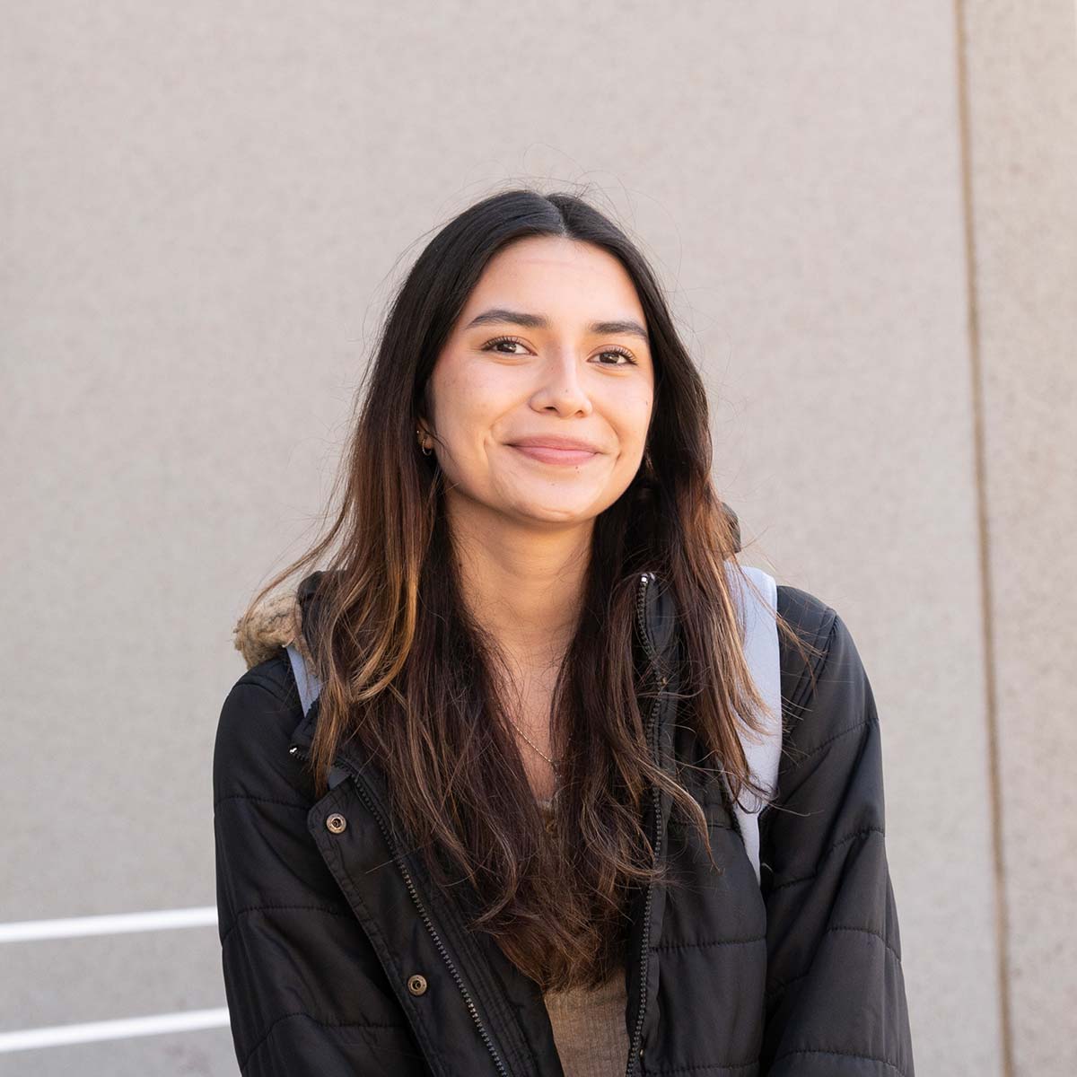 A young woman with long, wavy brown hair and light skin is smiling softly at the camera. She is wearing a black puffer jacket with a fur-lined hood and carrying a light-colored backpack. The background features a neutral beige wall with a white railing.