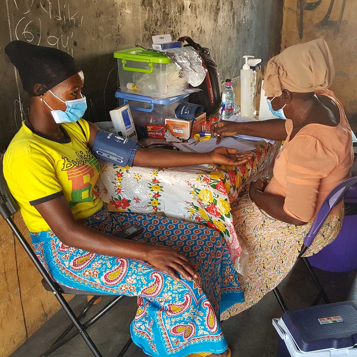 Two women sit facing each other at a table. The woman on the left wears a blood pressure cuff, while the woman on the right writes in her notebook.