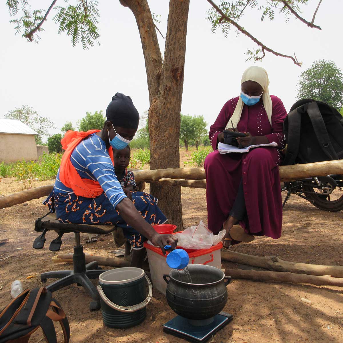 Two women sit under a tree around a cooking fire with pots. One holds an open notebook while the other pours water into one of the pots.