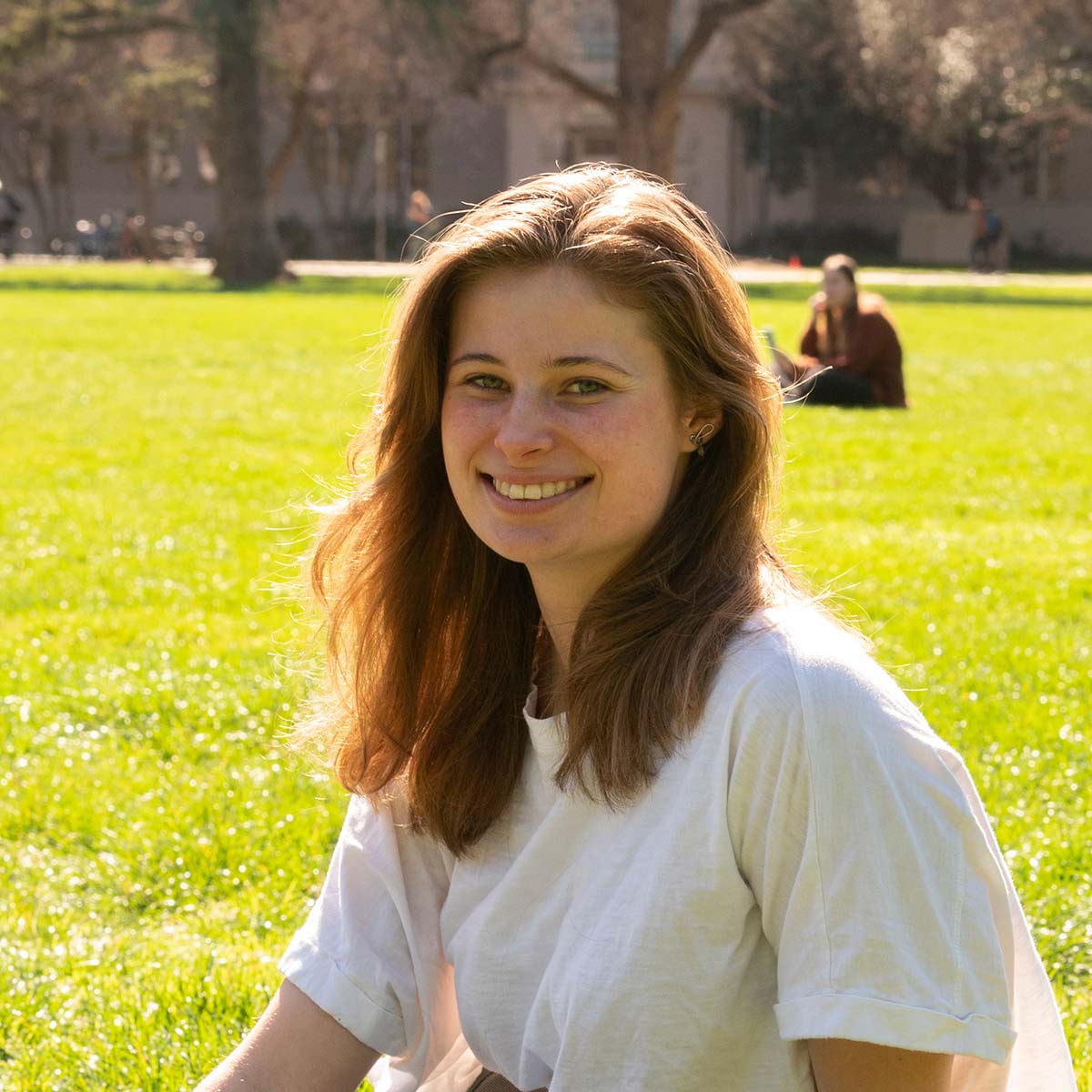 A young woman with shoulder-length, wavy light brown hair and light skin is smiling at the camera. She is wearing a loose-fitting white T-shirt. The background features a sunlit grassy field with people sitting in the distance.