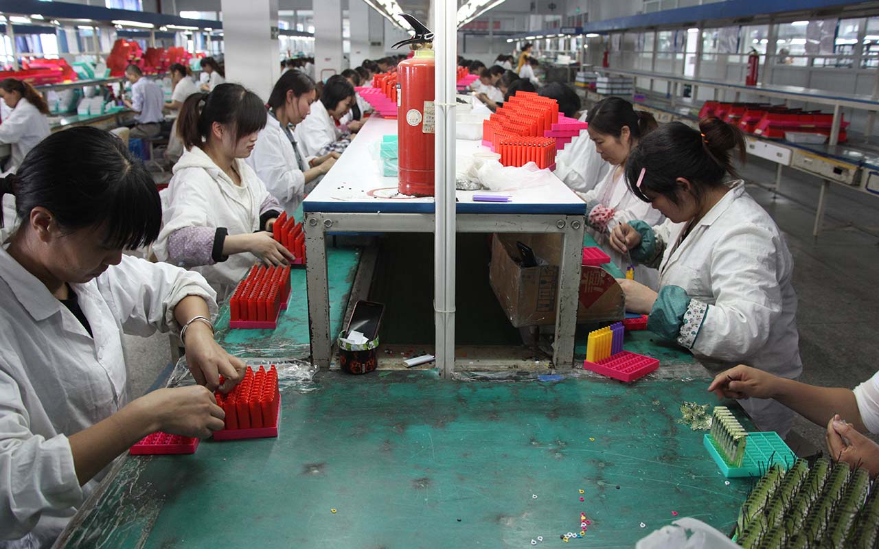 Photo of the inside of a factory. Two rows of workers wearing white coats are seated at a long green table and inspect small red tube-like pieces of plastic.