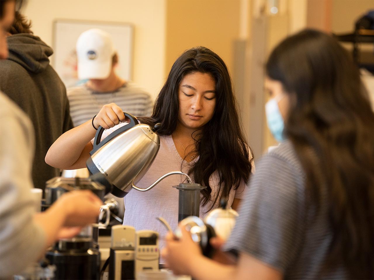 A student with long, dark hair pours boiling water from a stainless-steel kettle over coffee grounds in a dark cylinder. 