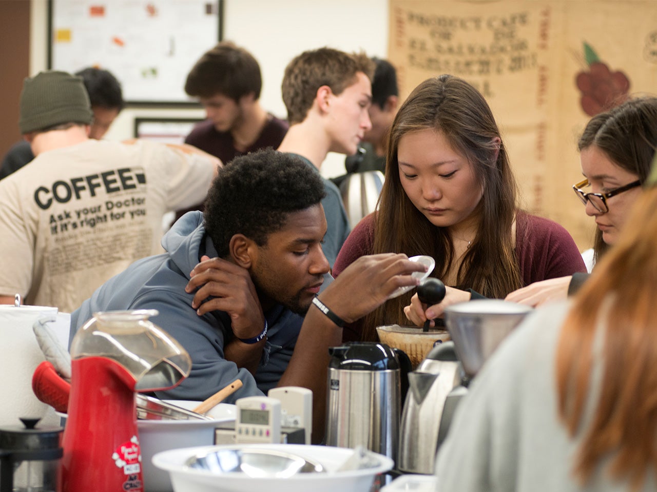 Two smiling students surrounded by coffee equipment in the foreground of the photo pour boiling water from a kettle over coffee grounds in a beaker while other students work on their lab projects in the background.