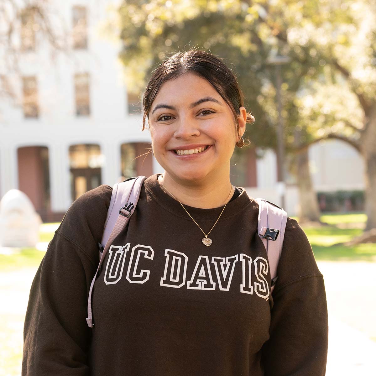 A young woman with dark hair tied back in a casual style is smiling at the camera. She is wearing a brown "UC Davis" sweatshirt, a gold heart-shaped necklace, and a light pink backpack. The background is slightly blurred, showing a sunny outdoor setting with trees.