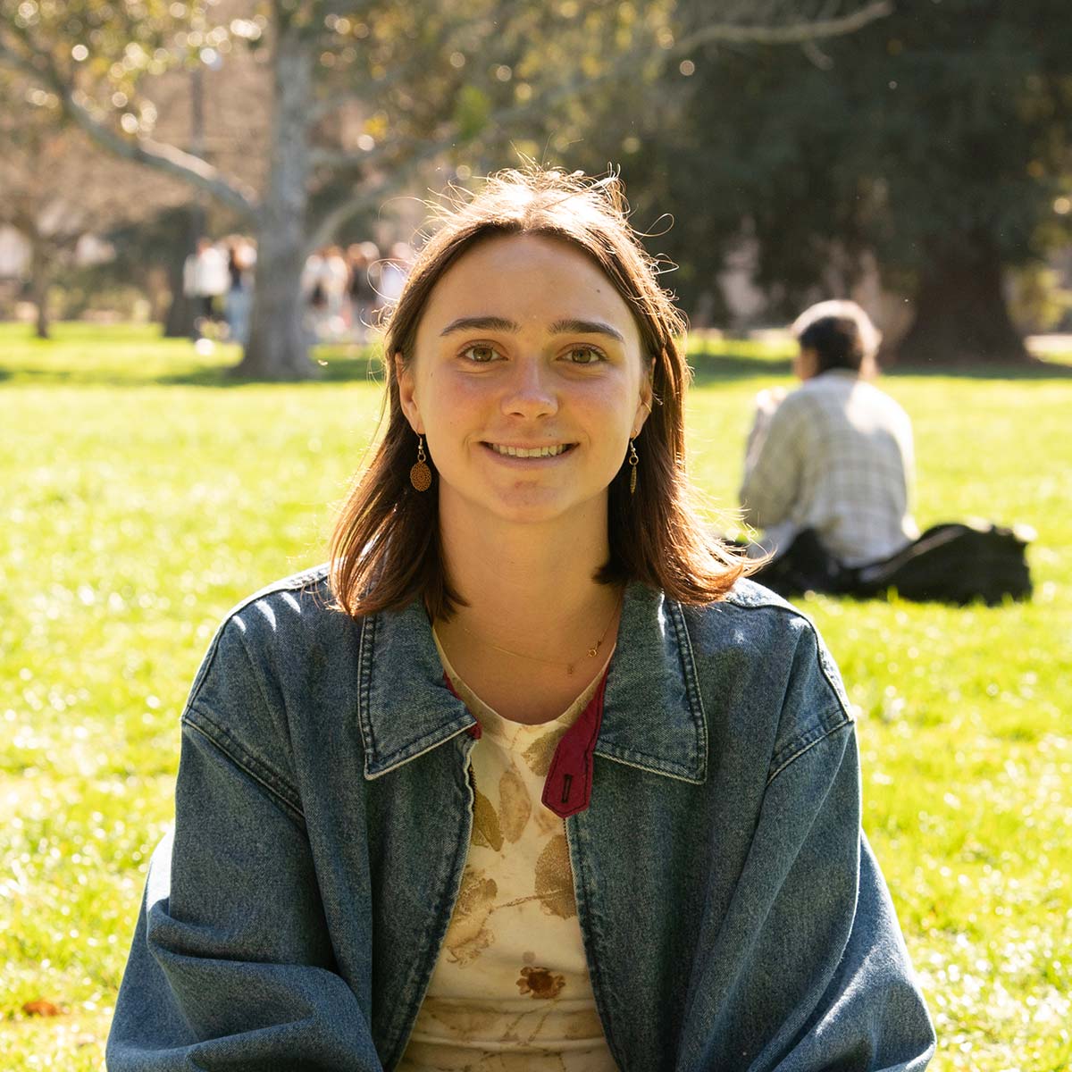 A young woman with shoulder-length brown hair and light skin is smiling at the camera. She is wearing a denim jacket over a floral-patterned top and gold dangling earrings. The background features a green grassy area with people sitting and walking, trees, and sunlight filtering through the leaves.