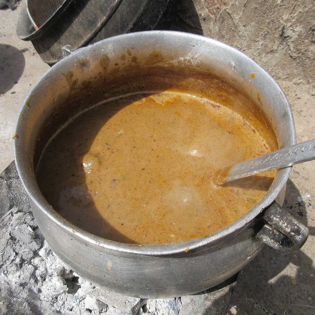 Photo of a silver-colored pot with a simmering brown broth in it. The pot is near the bare ground outside, with burned coals underneath it.