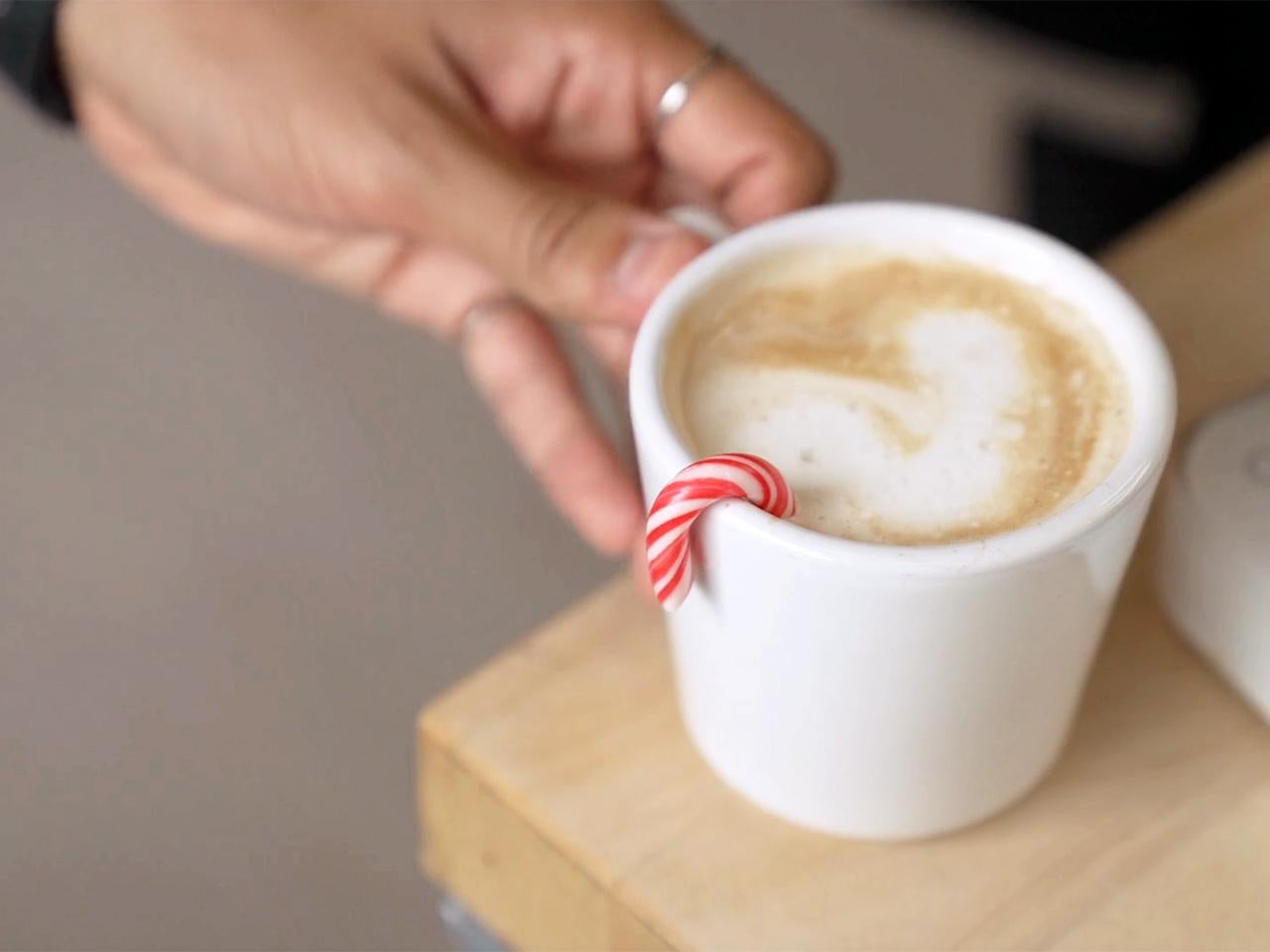 A closeup photo of a hand grabbing a small white mug filled with coffee and foam in the shape of a heart with a candy cane hanging on the rim for garnish.