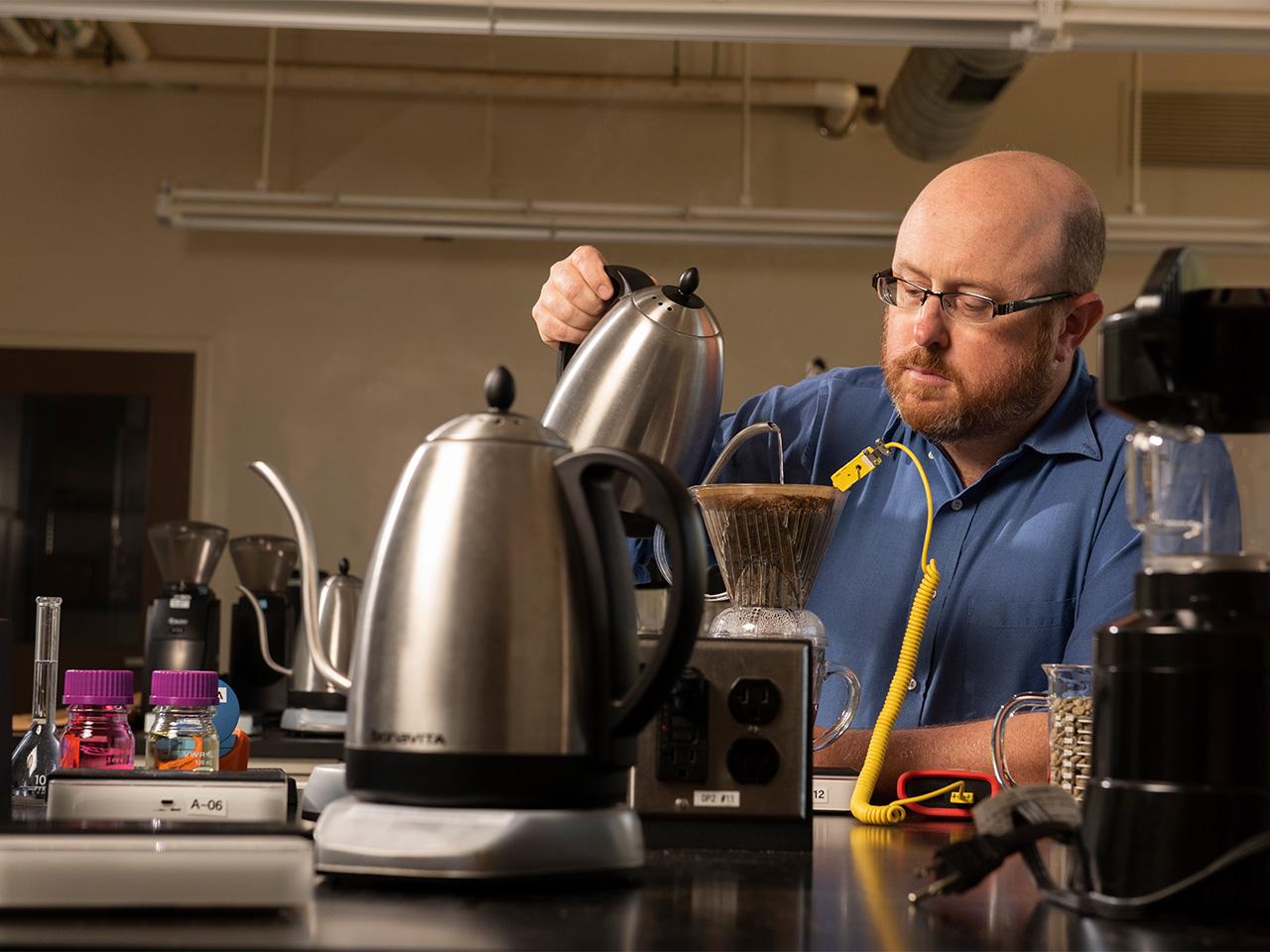 A bearded man in a blue button-down shirt and glasses concentrates while pouring hot water from a stainless-steel kettle over coffee grounds in a V60 plastic coffee dripper. He is surrounded by numerous scientific instruments and coffee tools.  