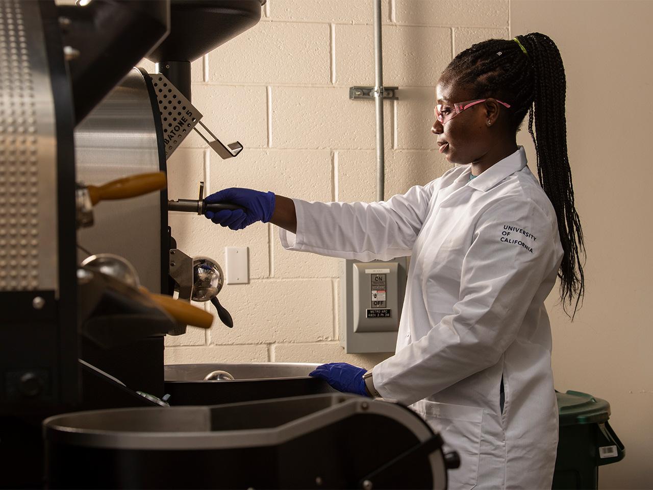 Laudia Anokye-Bempah, in full lab gear and a high ponytail, uses a stainless steel coffee roaster.