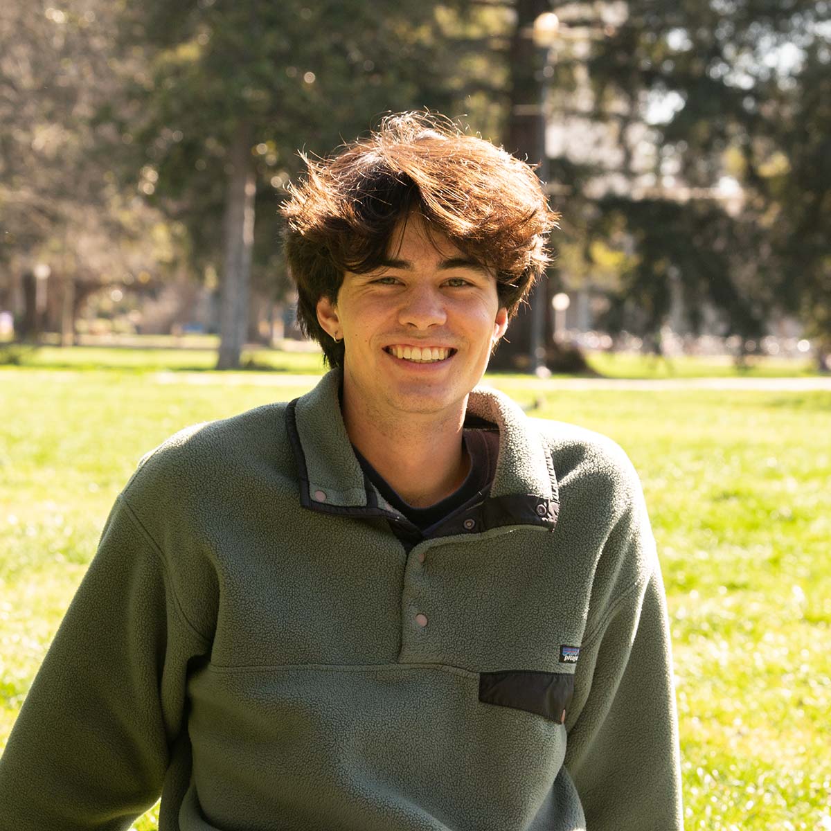 A young man with wavy brown hair and light skin is smiling at the camera. He is wearing a green Patagonia fleece pullover. The background features a sunlit grassy area with trees and a lamppost.