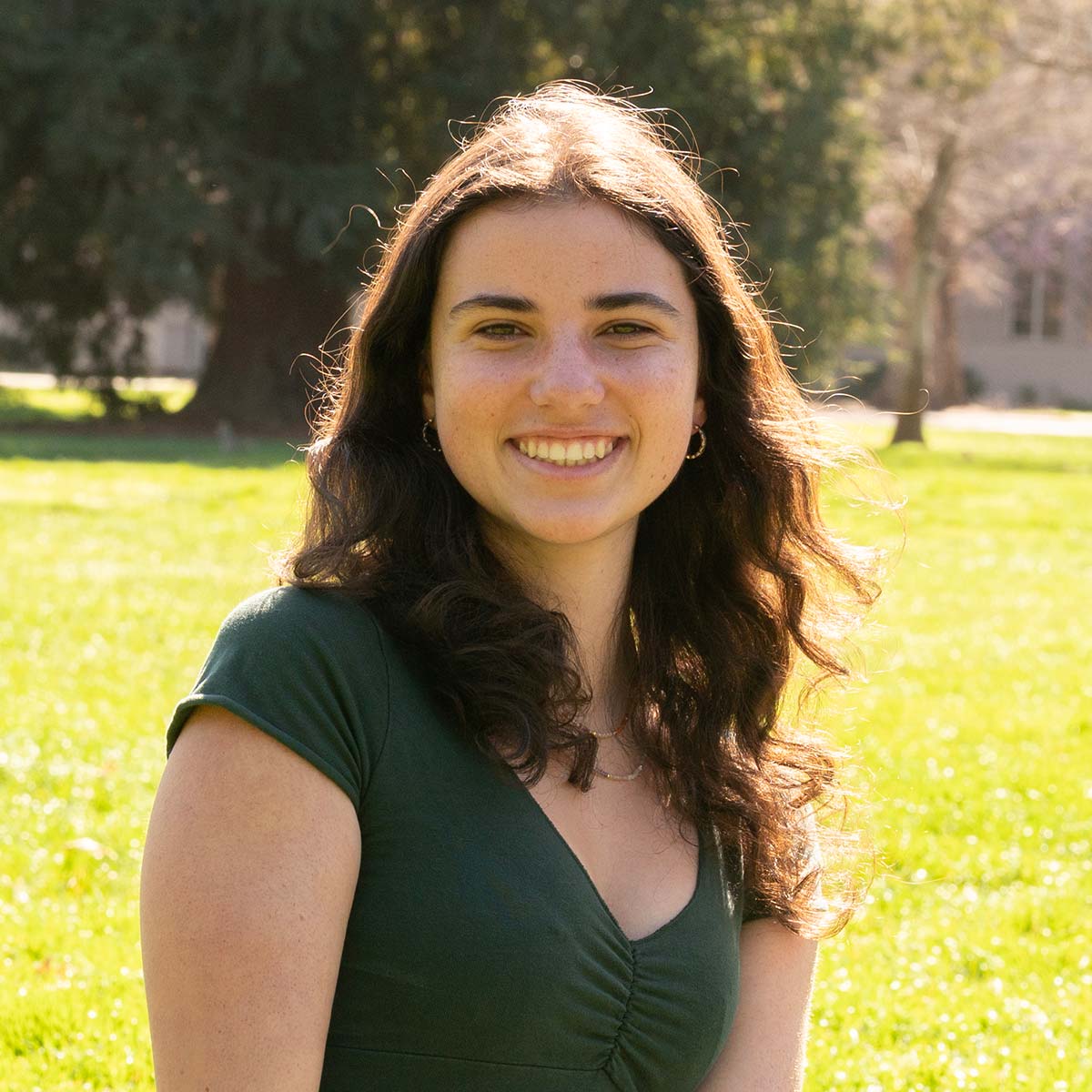 A young woman with long, wavy brown hair and light skin is smiling at the camera. She is wearing a dark green short-sleeved top with a V-neckline. The background features a sunlit grassy area with trees.