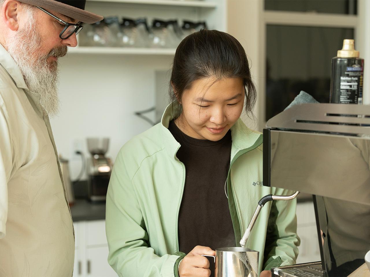 Timothy Styczynski, a man with a white beard, glasses, and light button-down shirt, watches as student Peyton Yang, wearing a black shirt and mint green jacket, uses an espresso machine to froth milk for a latte.