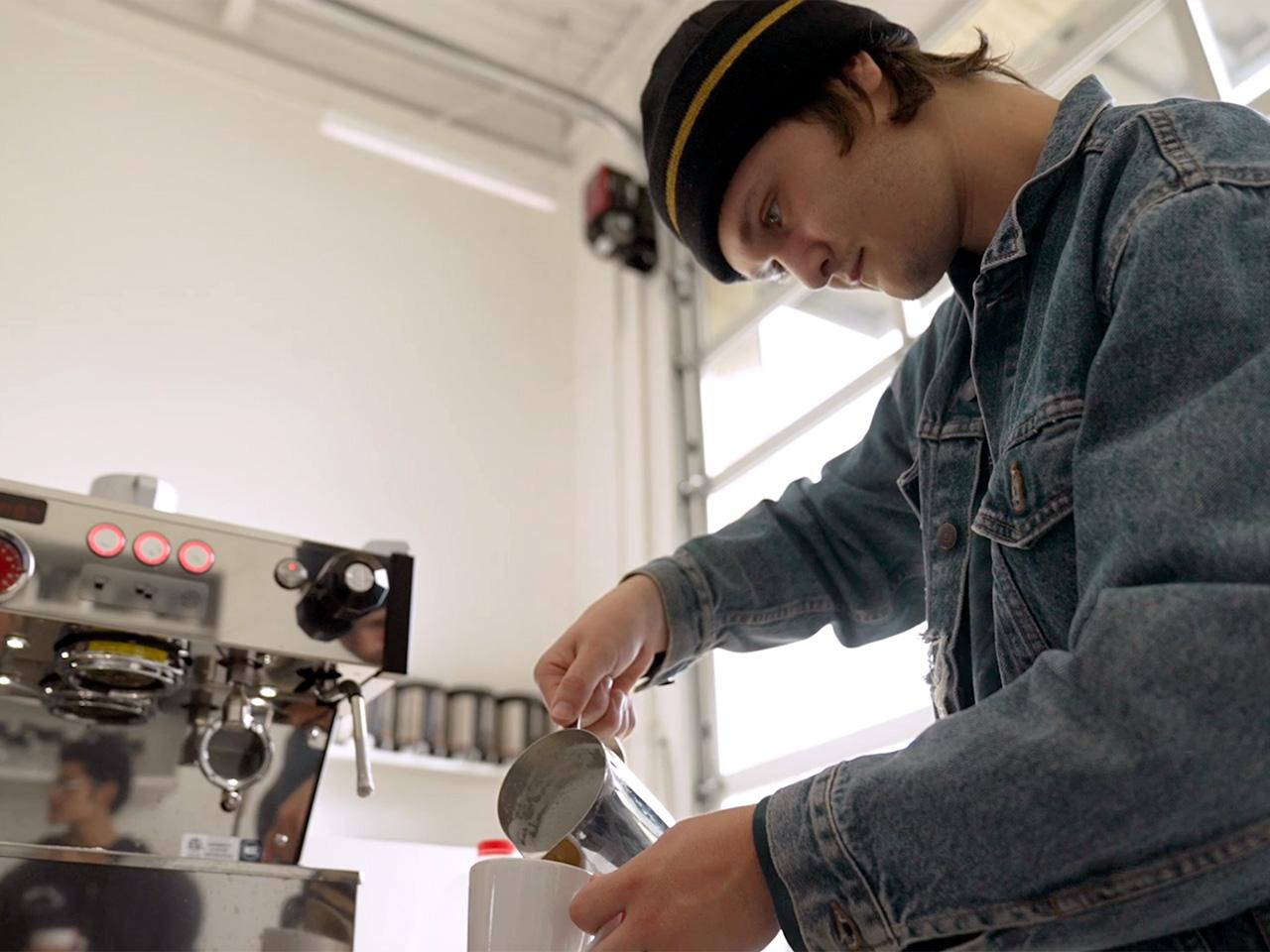 Student Keegan Thompson, wearing a dark beanie and blue denim jacket, pours foam into a white mug.