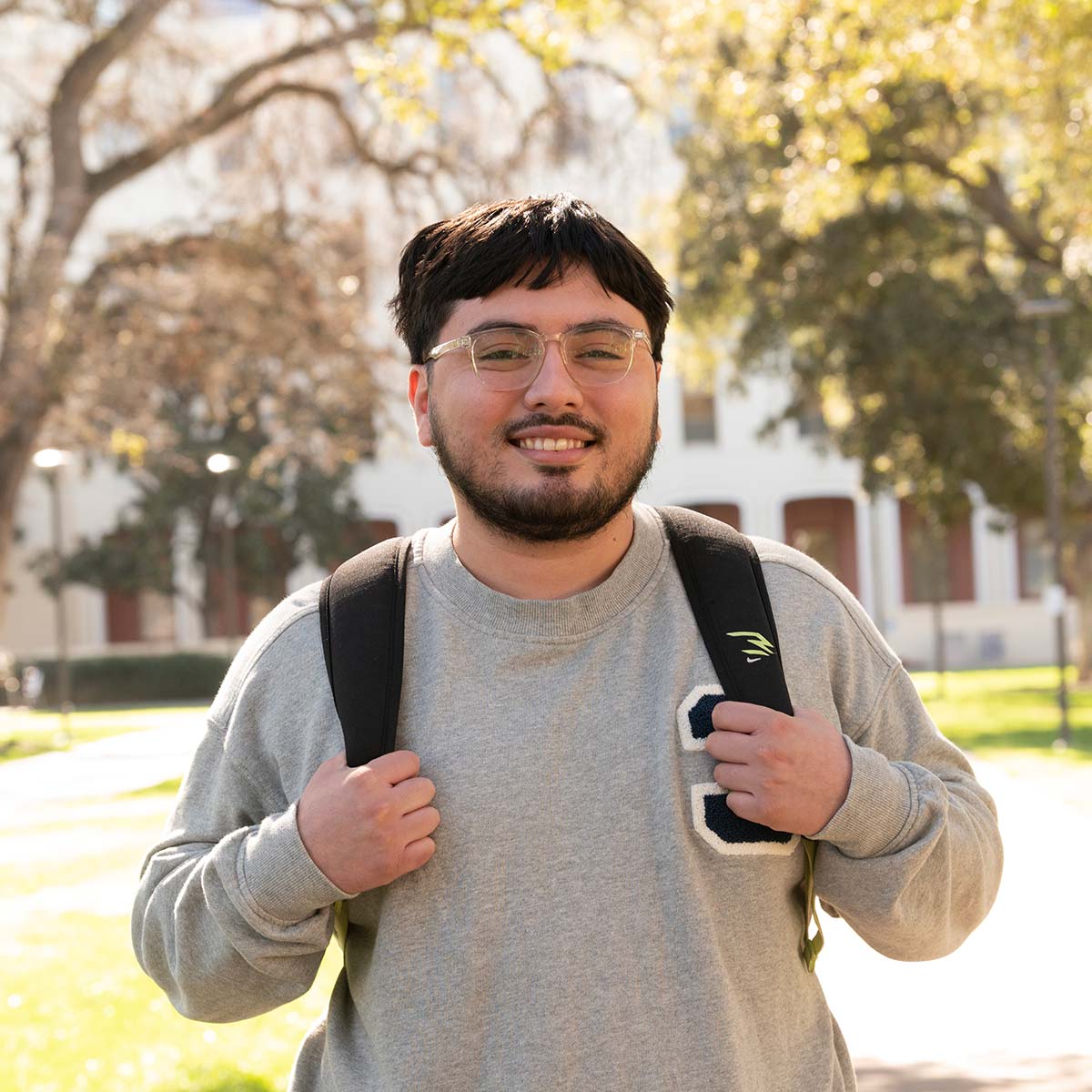 A young man with short black hair, a beard, and glasses is smiling at the camera. He is wearing a gray sweatshirt with a letter patch on the chest and carrying a black backpack. The background features a sunlit college campus with trees, green grass, and a white building with large windows.