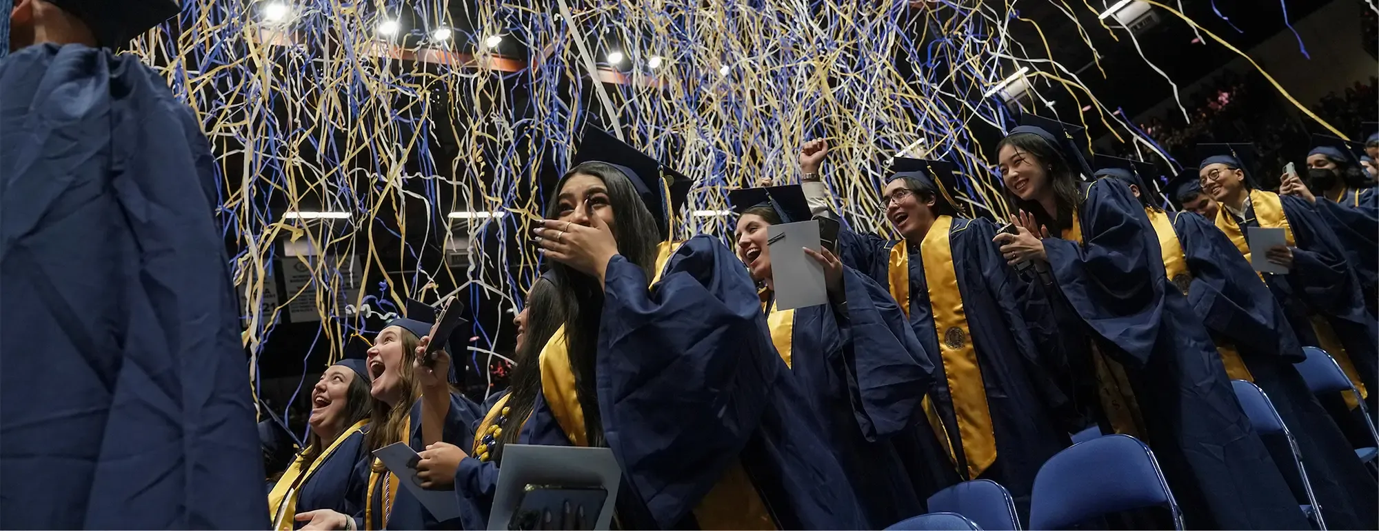 Graduates in navy gowns and gold stoles cheer as gold streamers fall