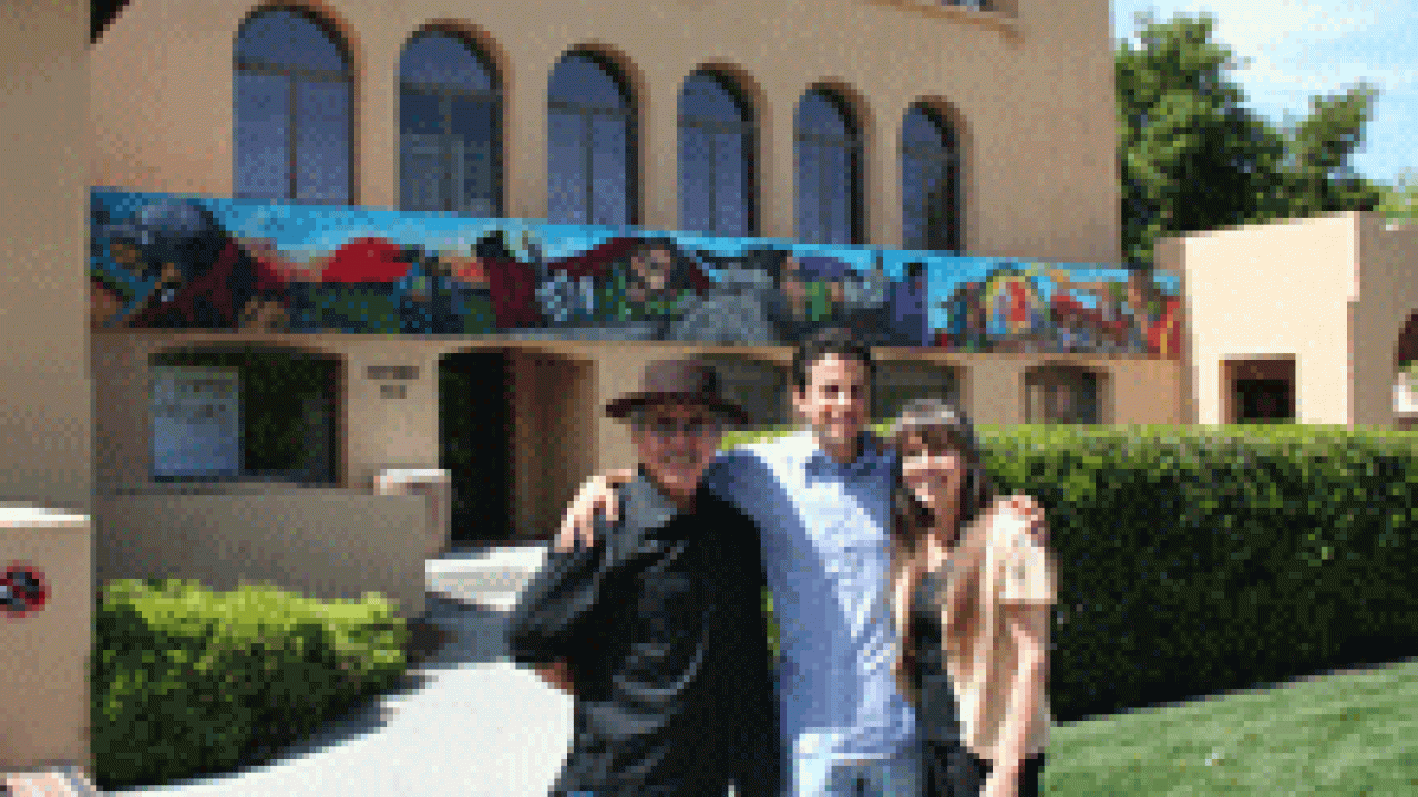 Chicana/o studies and art professor Malaquias Montoya, left,  painted this mural on the outside of the El Centro Chicano building at Stanford in 1981. He is pictured with his wife, Lezlie Salkowitz-Montoya, right, and son Maceo, an artist as wel