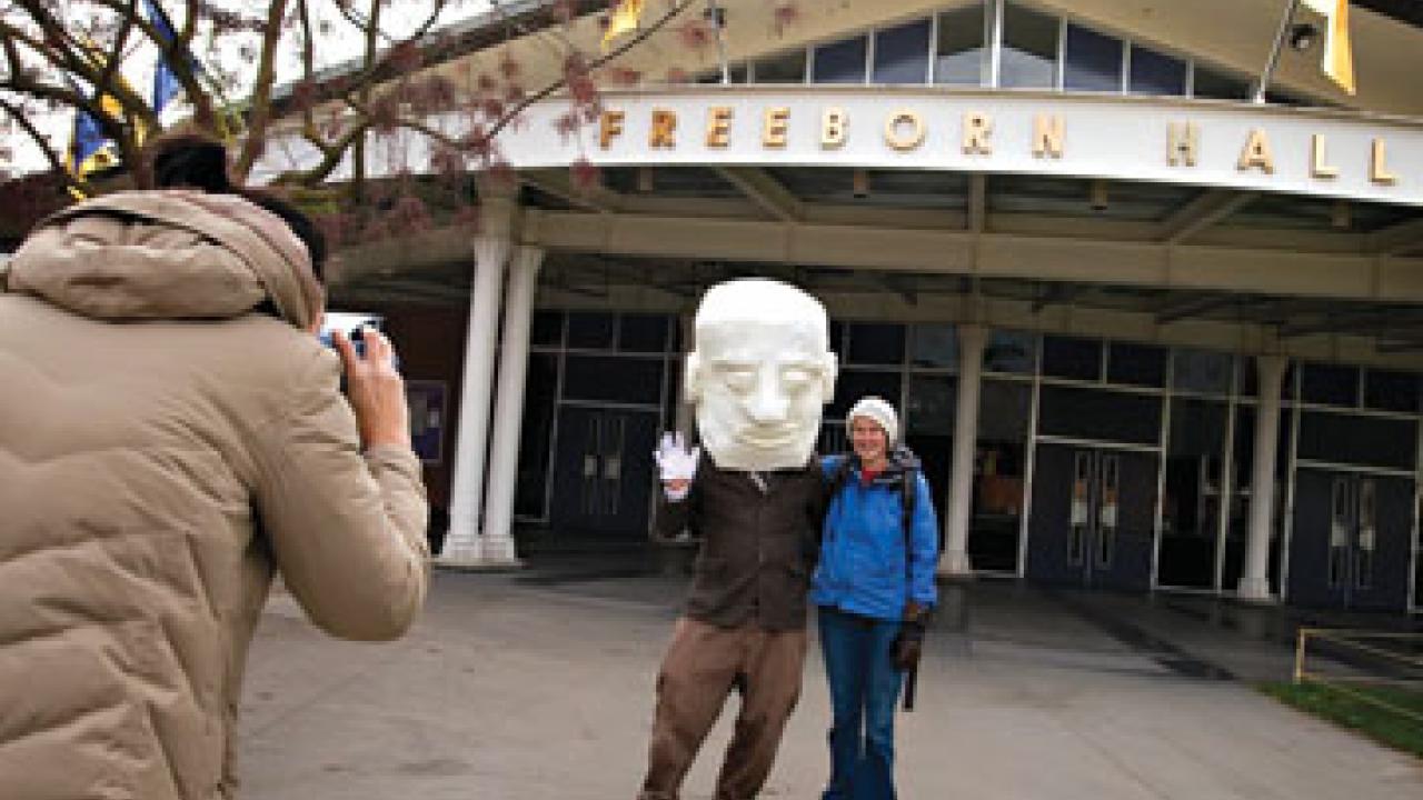 Ryan Bulis wears the Freeborn mask, as Anna Ng takes a Polaroid picture of "Freeborn" and an unidentified passer-by in front of Freeborn's namesake ha