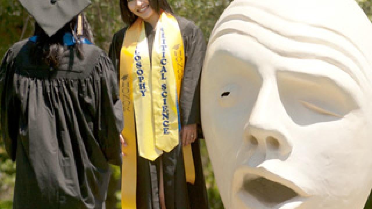 Photo: Two women in commencement gowns and mortar board hats near an Egghead sculpture