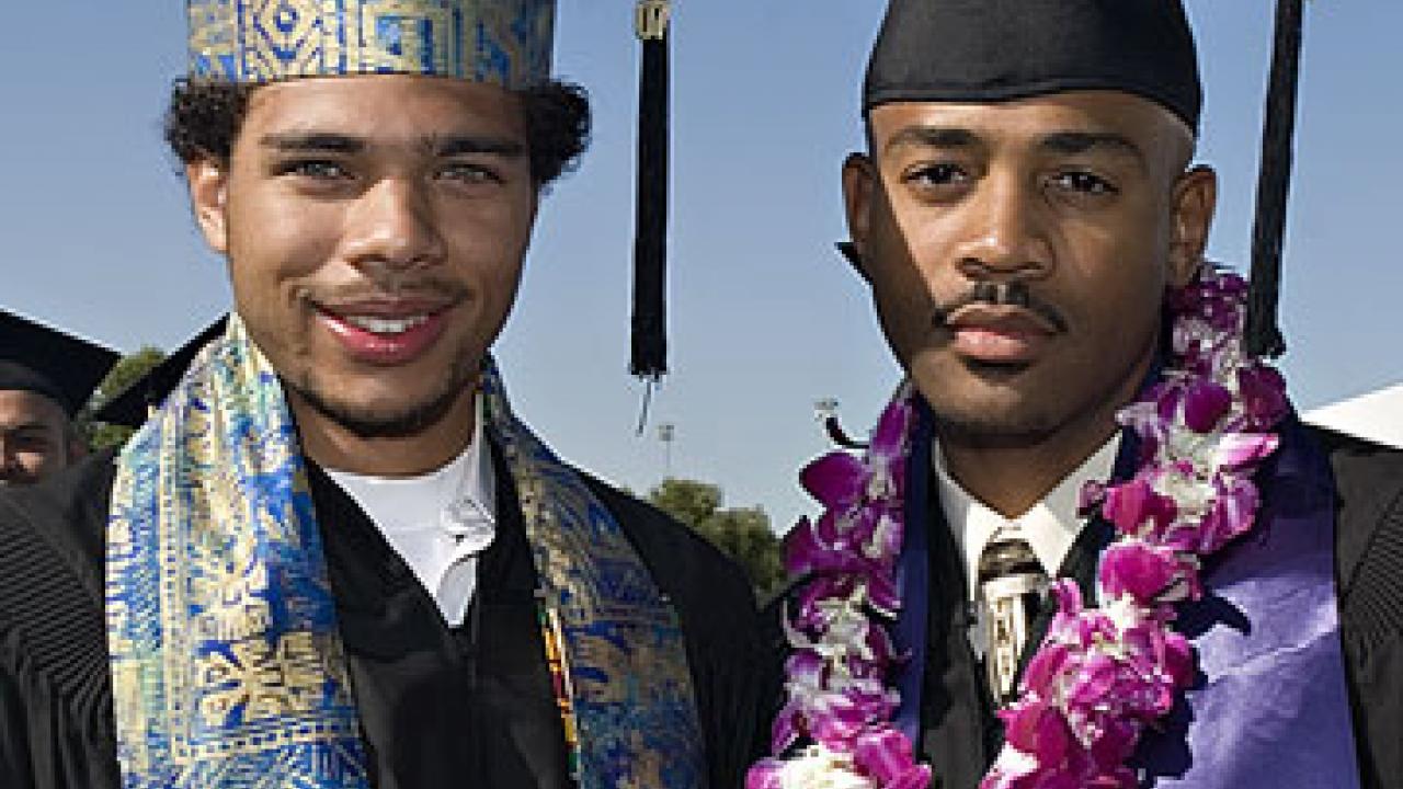 Photo: two graduates, one in colorful mortar board, the other with a purple lei