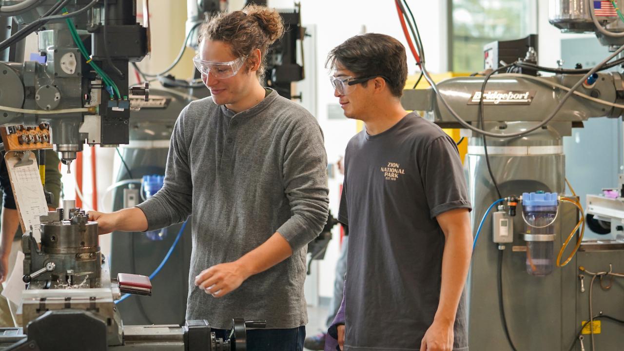 A male student wearing a grey t-shirt and clear protective glasses operates machinery in a student workship, while a male student wearing a blue t-shirt and clear protective goggles stands to the right and looks onward at the machinery.  