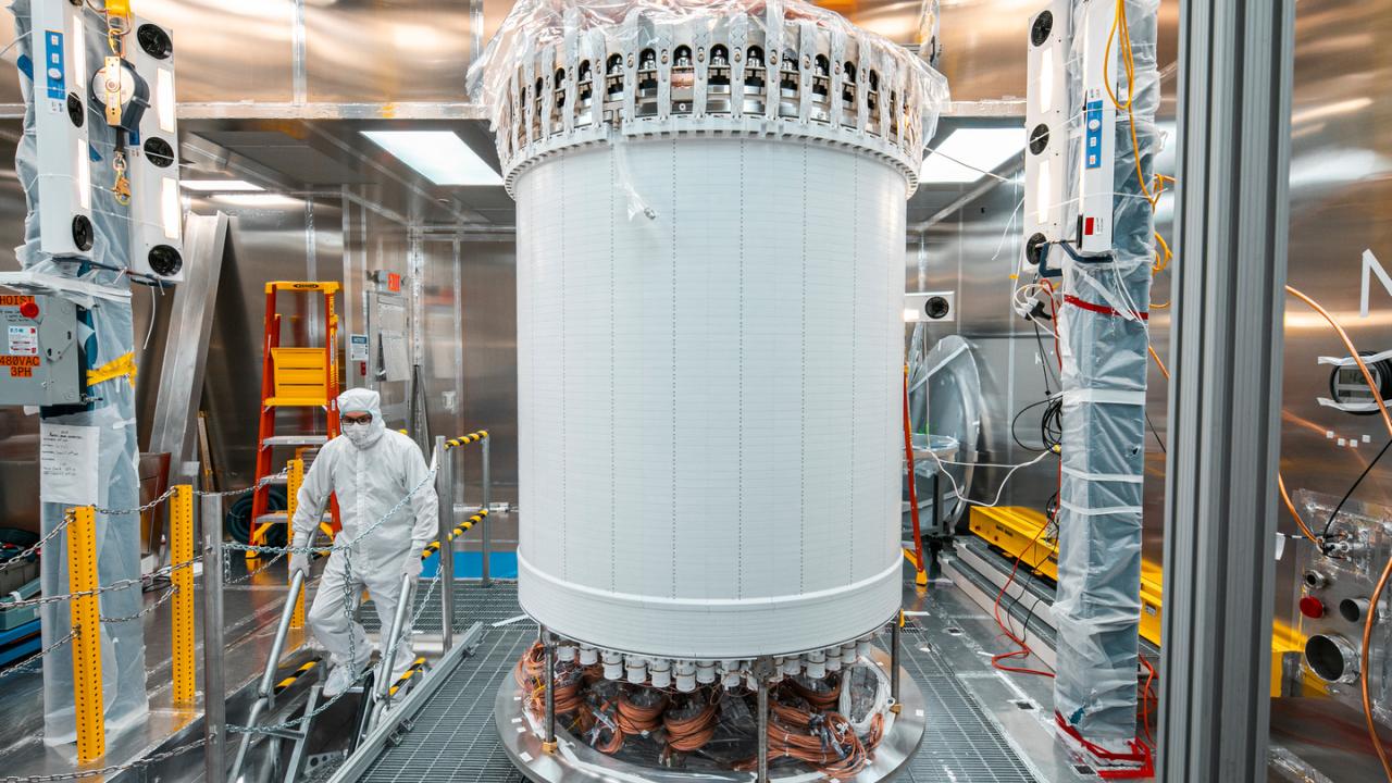 A large white cylinder in the middle of a laboratory setting. To the left a person in a white clean room coverall and mask descends a staircase. 