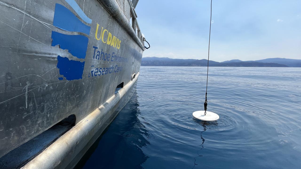 White secchi disc hanging from cord off research vessel labeled UC Davis Tahoe Environmental Research Center hovers over surface of blue Lake Tahoe