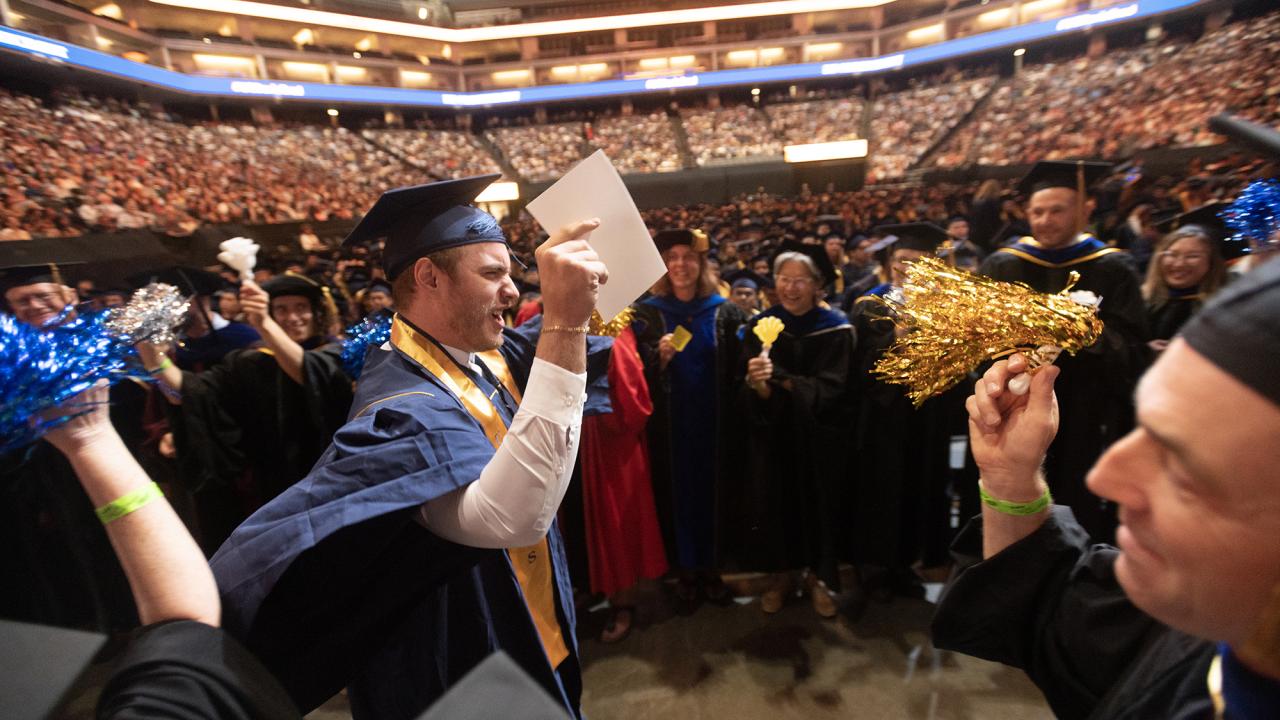 With a hand in the air, a male graduate strides between two lines of faculty members shaking pompoms and clappers. 
