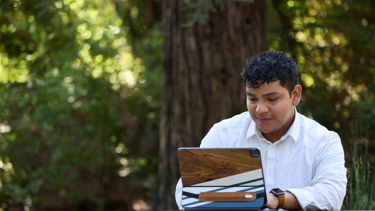 A student in a white button-down shirt, works on their iPad outside at a table in the UC Davis Redwood Grove.  