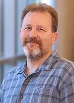 inside a university building a man with a goatee, short hair and a grey flannel shirt smiles towards the camera