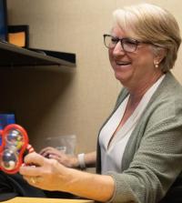 a woman with black rimmed glasses and white hair holds a child's toy while inside a medical facility, she is smiling towards her patient.