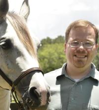 a man wearing glasses and a green shirt stands to the left of a white horse, who takes up the left side of the photo. they're outside in a stable setting.