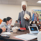 Chancellor Gary S. May, standing, talking, at a table full of students.