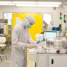 Person in a white clean room suit with hood, eye protection and mask working at a bench in a high-tech setting. 