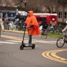 a cyclist in black tee and tan jacket, a man in orange parka and scooter and cyclist in helmet ride around a traffic circle with red Unitrans bus and cars in background