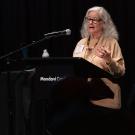 Standing a podium and illuminated by a stage light, Dr. Arline T. Geronimus speaks while staring at notes. She has long blonde hair past her shoulders, a white blouse and glasses with a red frame. 